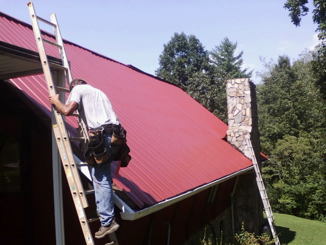 Roofer on a ladder beside a red metal roof and stone chimney — Soffit & Fascia Repair by Roofing Brothers in Ocklawaha